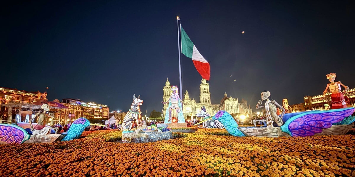 The Día de Muertos mega-ofrenda in the Zócalo of Mexico City at night, with giant skeleton figures, a carpet of marigolds, the Mexican flag, and the Metropolitan Cathedral