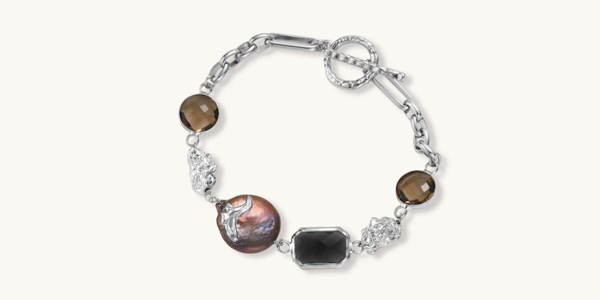 The Ofrenda Bracelet from the Courage Collection, a silver chain with a bronze baroque pearl, smoky quartz, and a black faceted stone, on a cream background