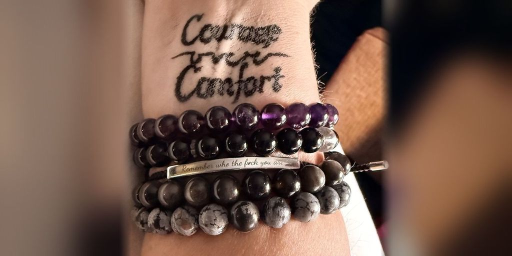 Carrie's wrist with her Courage over Comfort tattoo above a stack of mala bead bracelets, including a silver bar engraved Remember who the f*ck you are. Carrie's own photo
