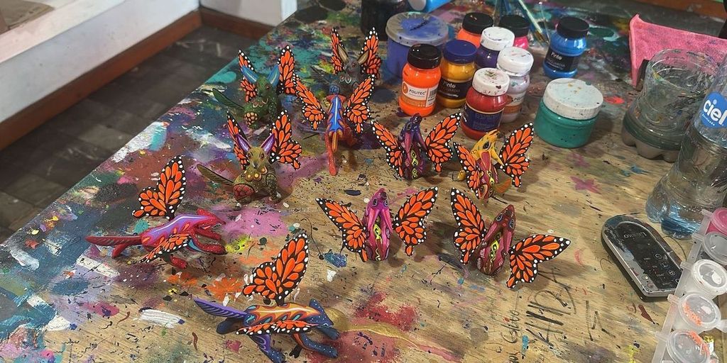 Hand-carved alebrijes with monarch wings on a paint-splattered worktable beside jars of paint, in an alebrije workshop in Oaxaca.